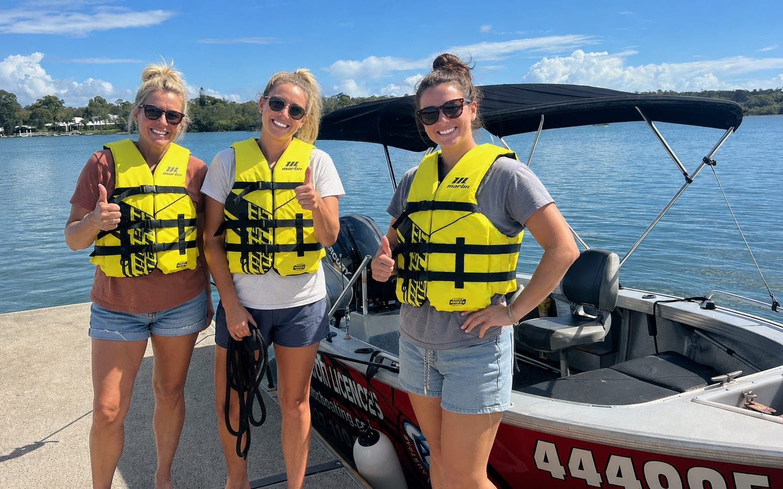 Three students in lifejackets giving a thumbs up beside the ABC Boating training boat after their Queensland boat licence course