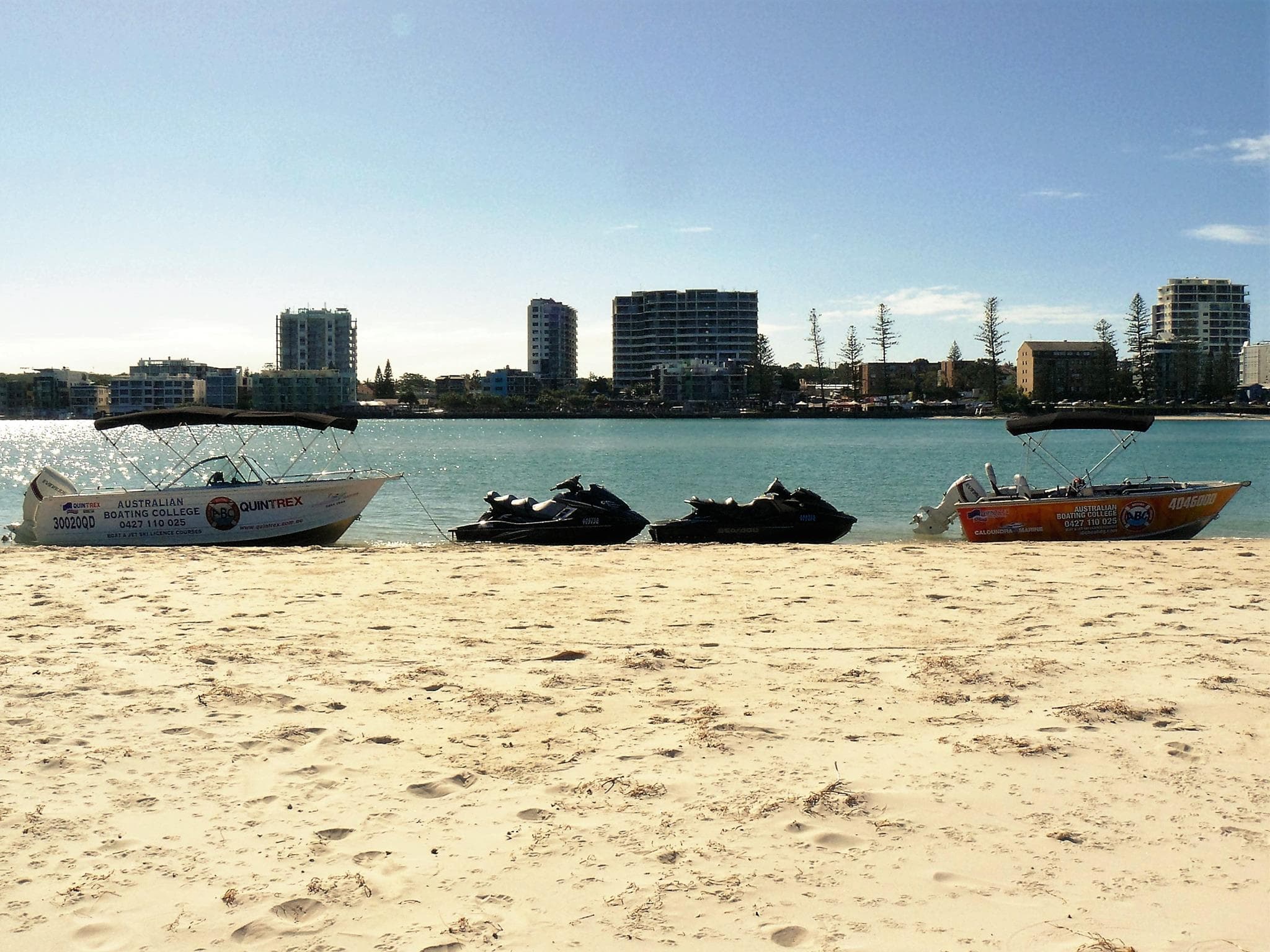 Training boats and jet skis on a sandy Pumicestone Passage beach with Caloundra's waterfront towers across the water