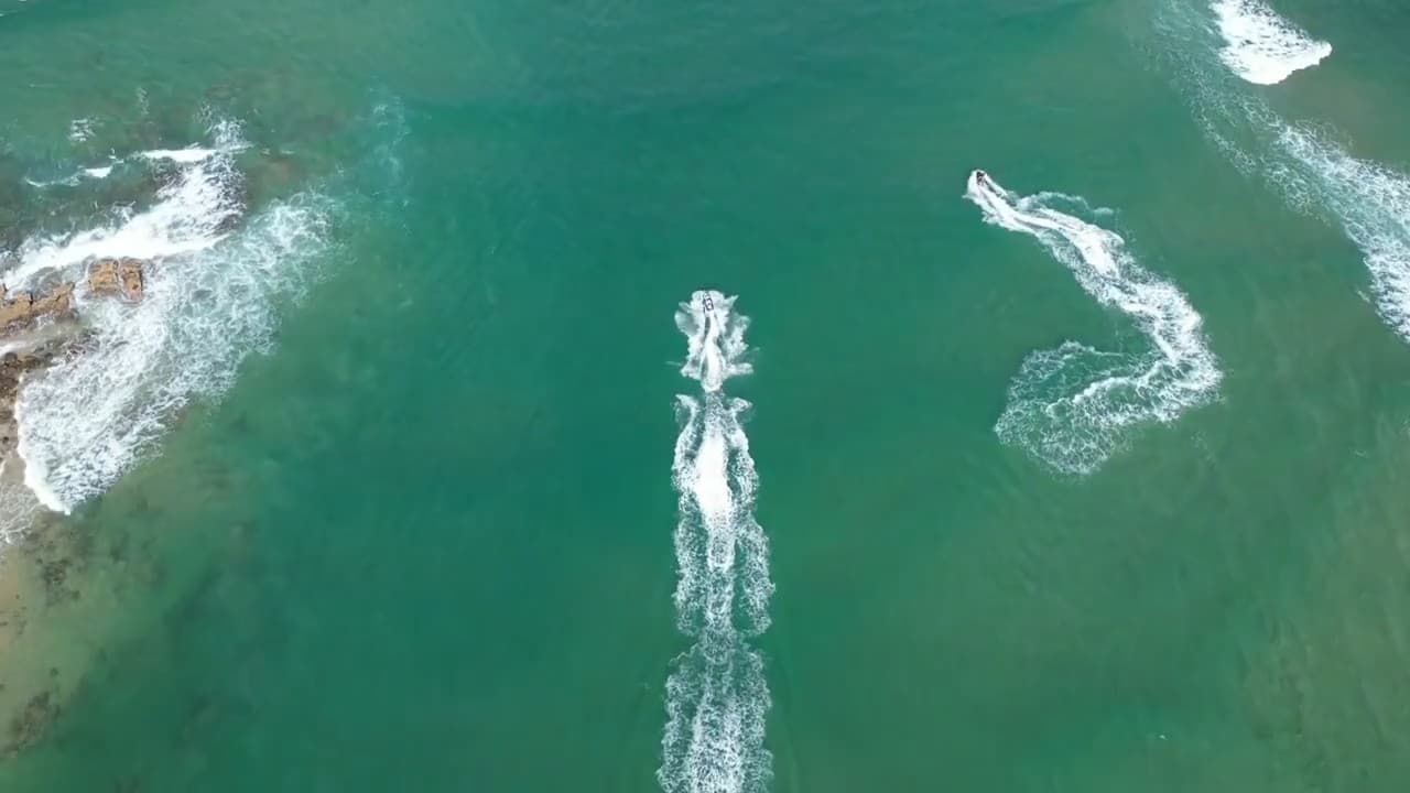 Overhead aerial view of two jet skis crossing clear turquoise water off a rocky Sunshine Coast headland, their wakes trailing behind them