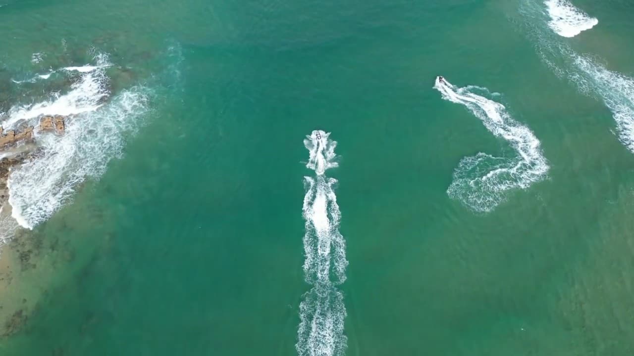 Overhead aerial view of two jet skis crossing clear turquoise water off a rocky Sunshine Coast headland, their wakes trailing behind them